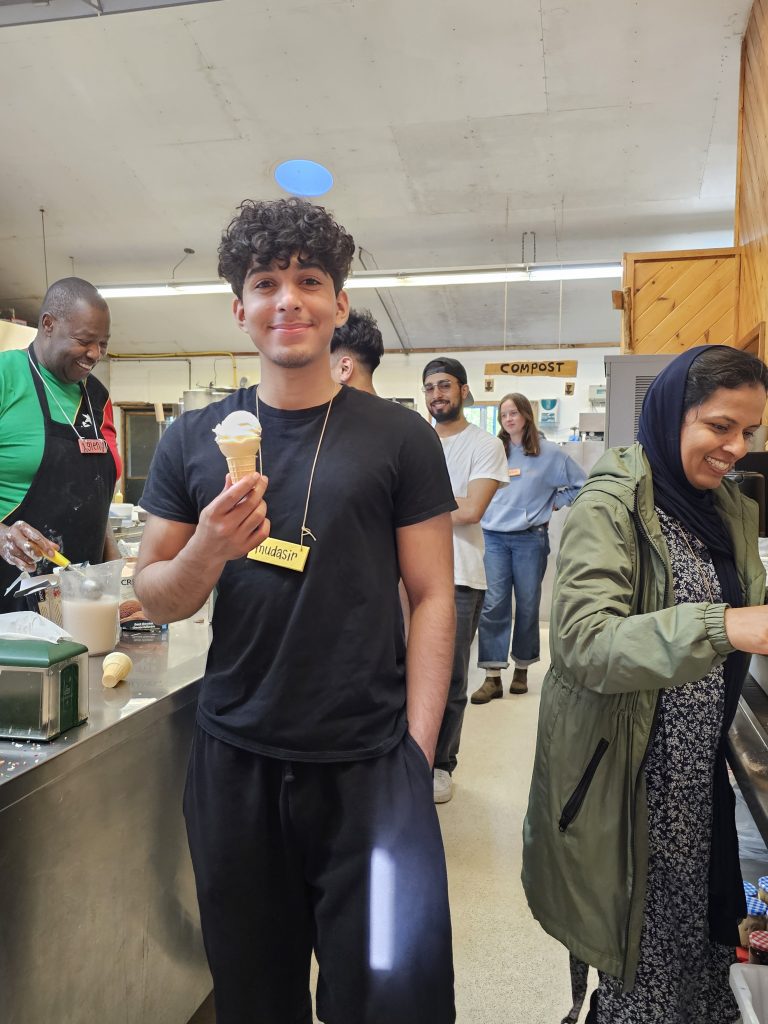 A young person with a name tag reading Mudasir stands with an ice cream cone in a cafeteria