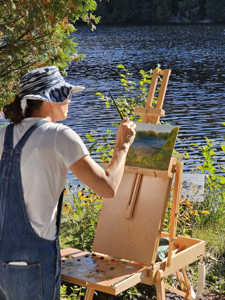 A person in a floppy hat works on a painting by the waterside