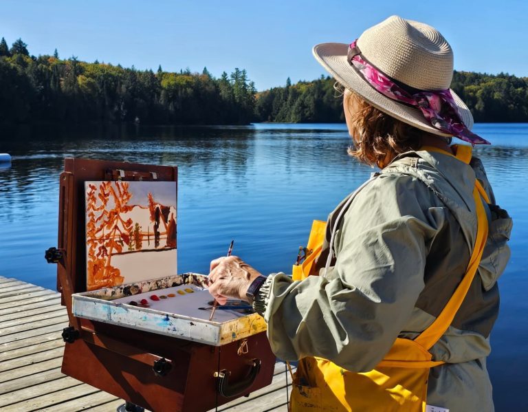 A woman on a dock looks at the landscape while standing in front of an art easel