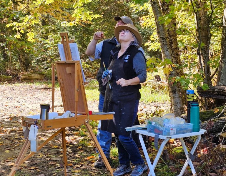 Two people under a tree and in front of an easel look upward