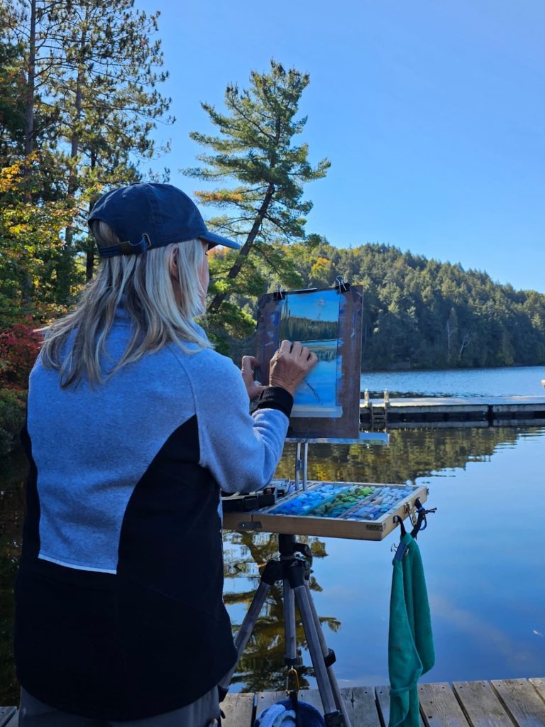 A woman in a sweater works on a lakeside landscape painting