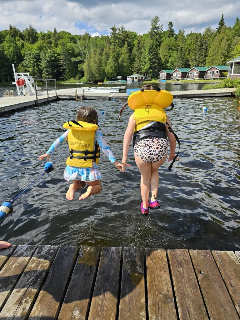 Two kids in life jackets leap from a dock