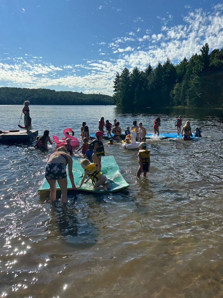 A large group of children and adults swim in a lake and play with a floating mat