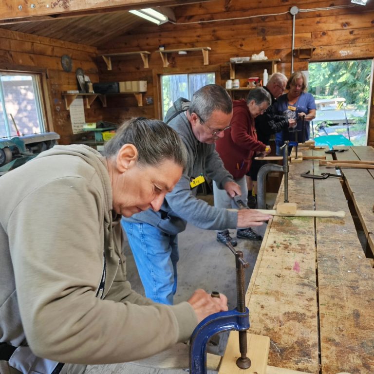 Woodworkers in a row, performing some sort of task with pieces of wood clamped to a table