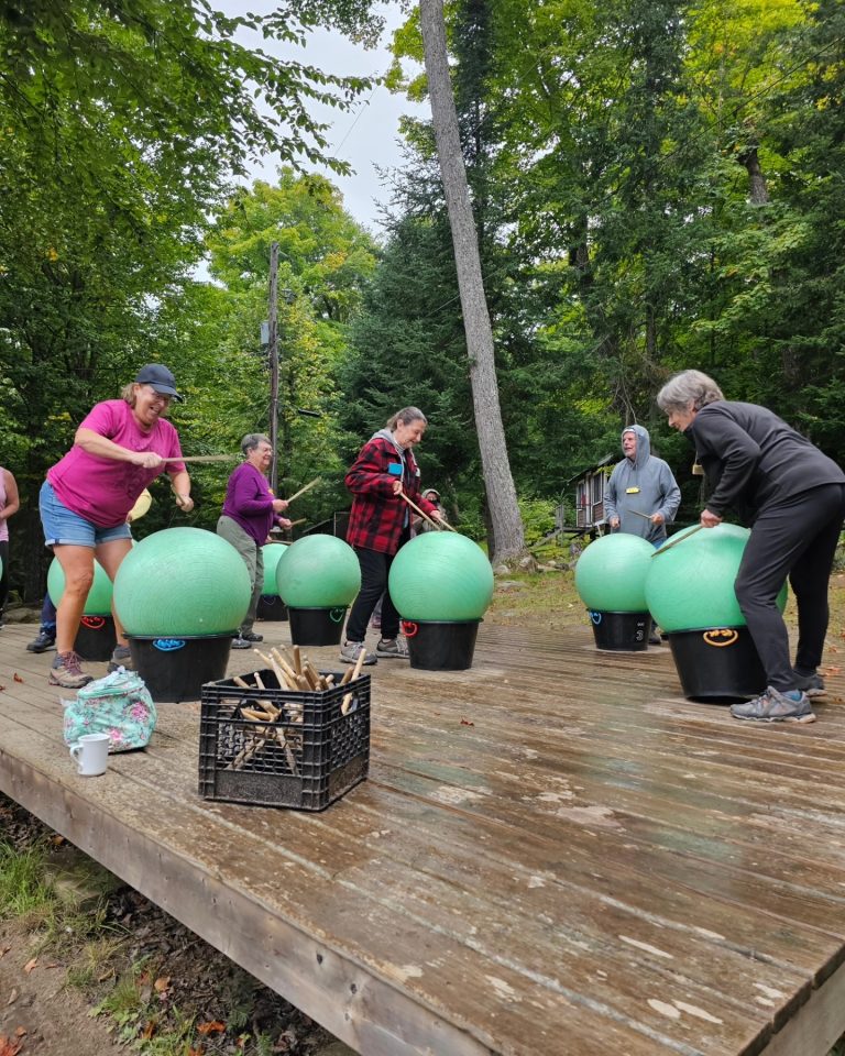 A group of people does some sort of outdoor exercise involving drumming on yoga balls in milk crates