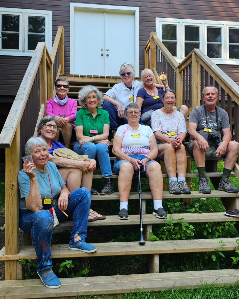 A group of nine smiling older people sit on the steps of a cabin