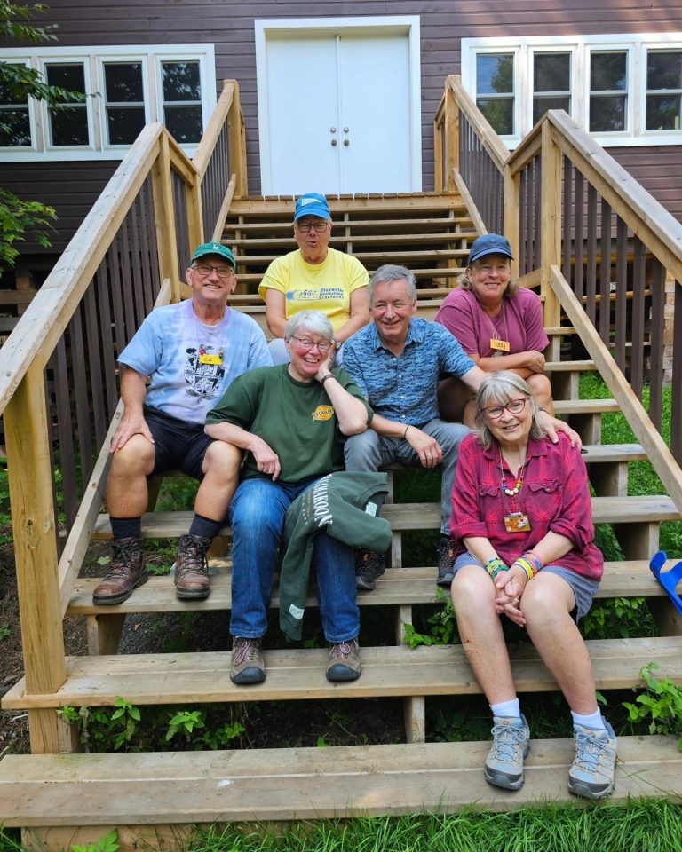 A group of six smiling older people sit on the steps of a cabin