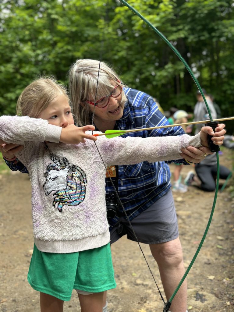 An older woman coaches a girl who is drawing back a bowstring
