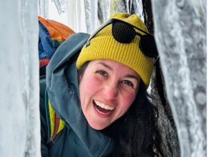 Inside an ice cave, a woman wearing a yellow jacket and blue hat smiles brightly at the camera.