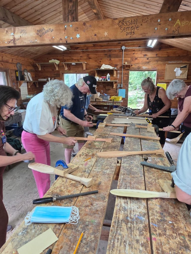 A group of people at a long work bench shaving canoe paddle blanks