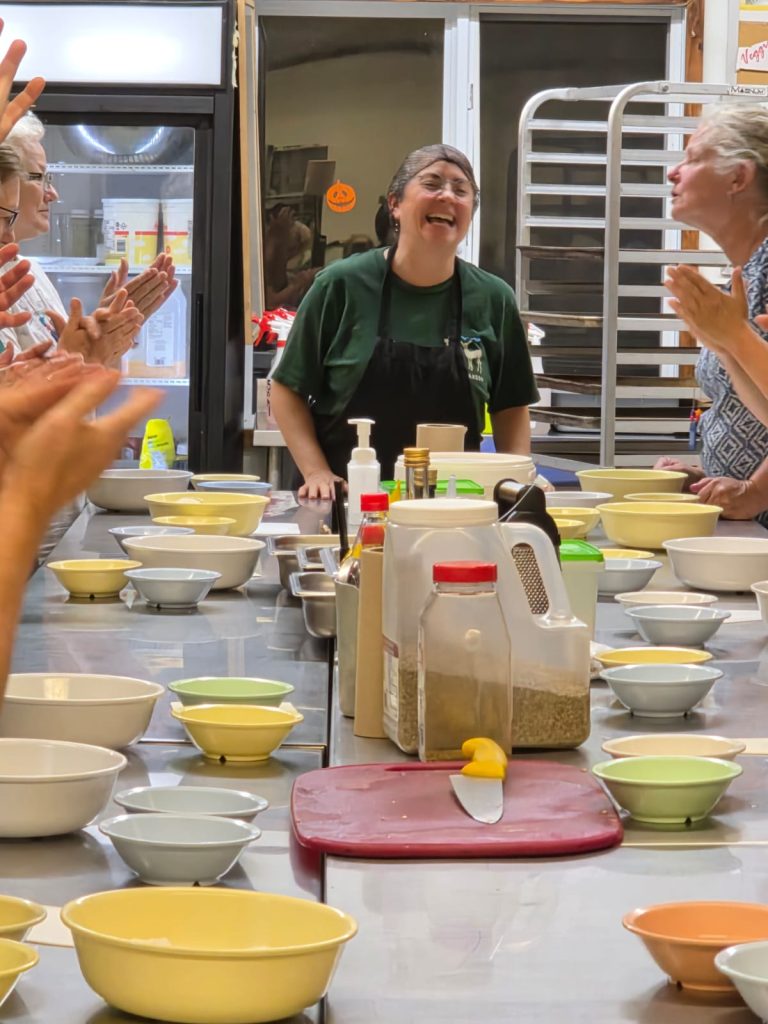 A laughing person in an apron stands at the head of a cooking class