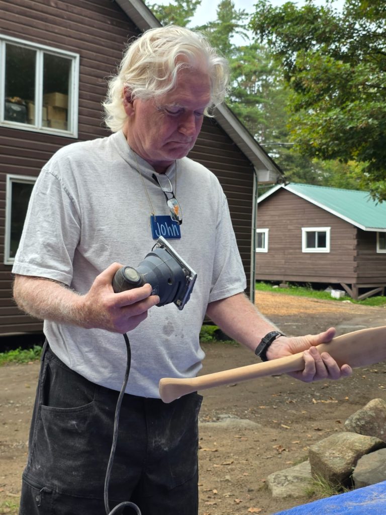 A man holding a palm sander inspects a small canoe paddle