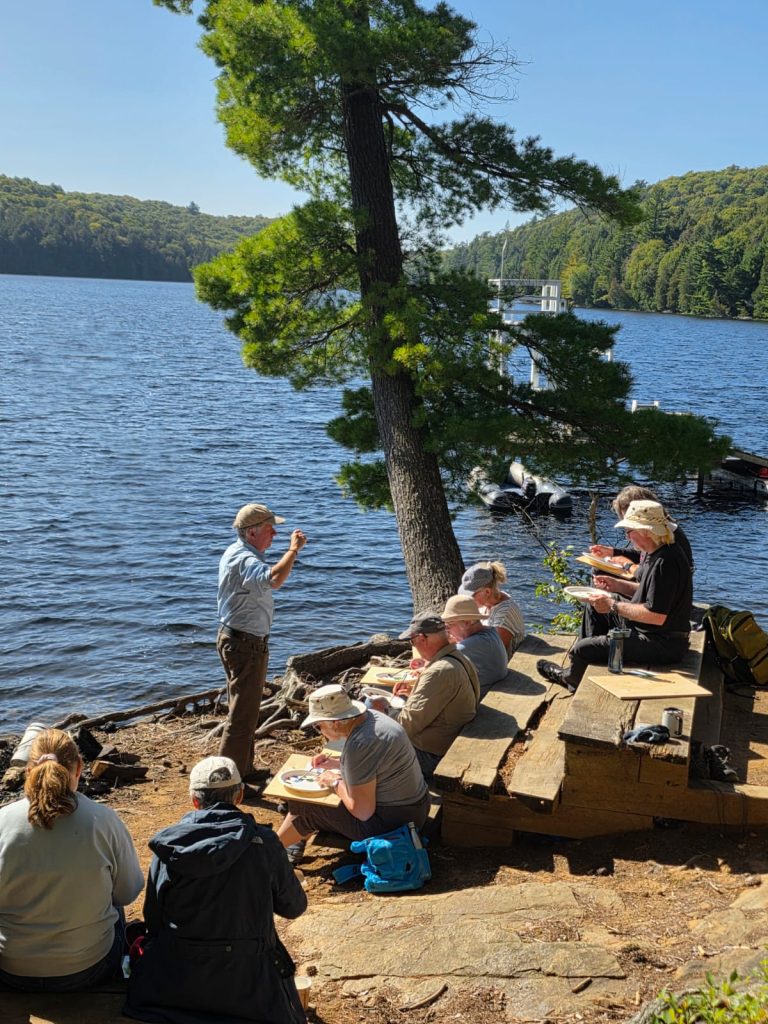 A group painting class sits on benches beside a lake