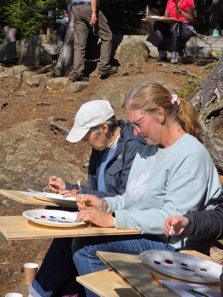 Two people with paint palettes sit outdoors working on paintings