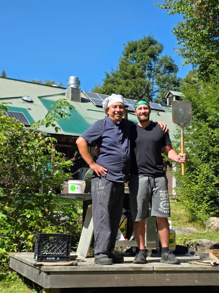 A man in a chef's hat and a man in a bandana stand together, with one raising a pizza paddle