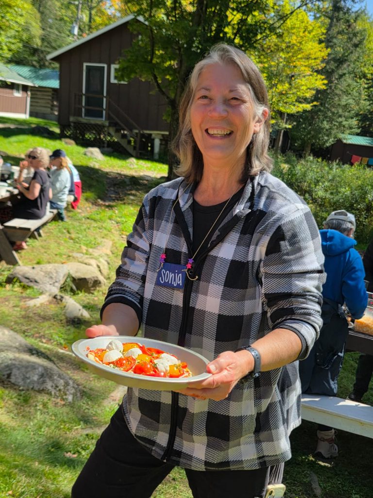A smiling woman stands outdoors with a plate of fresh tomatoes and cheese