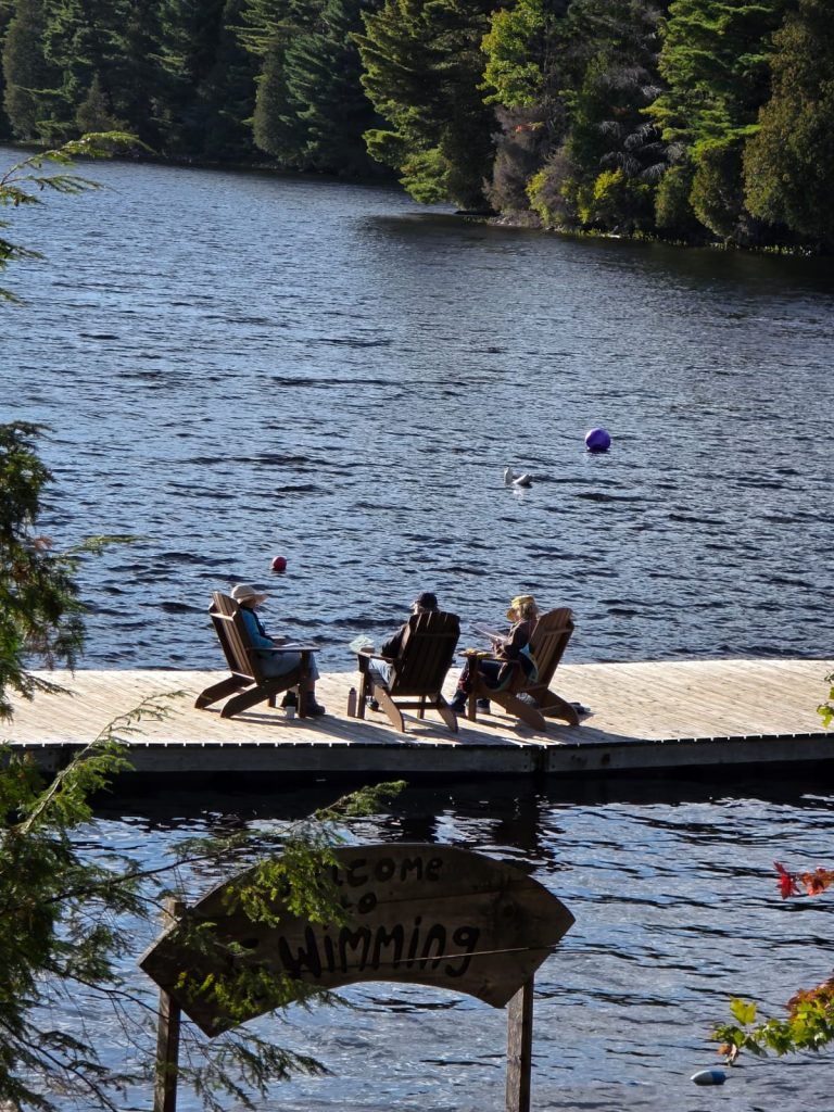 Three people sit in Muskoka chairs on a dock, seen from a distance
