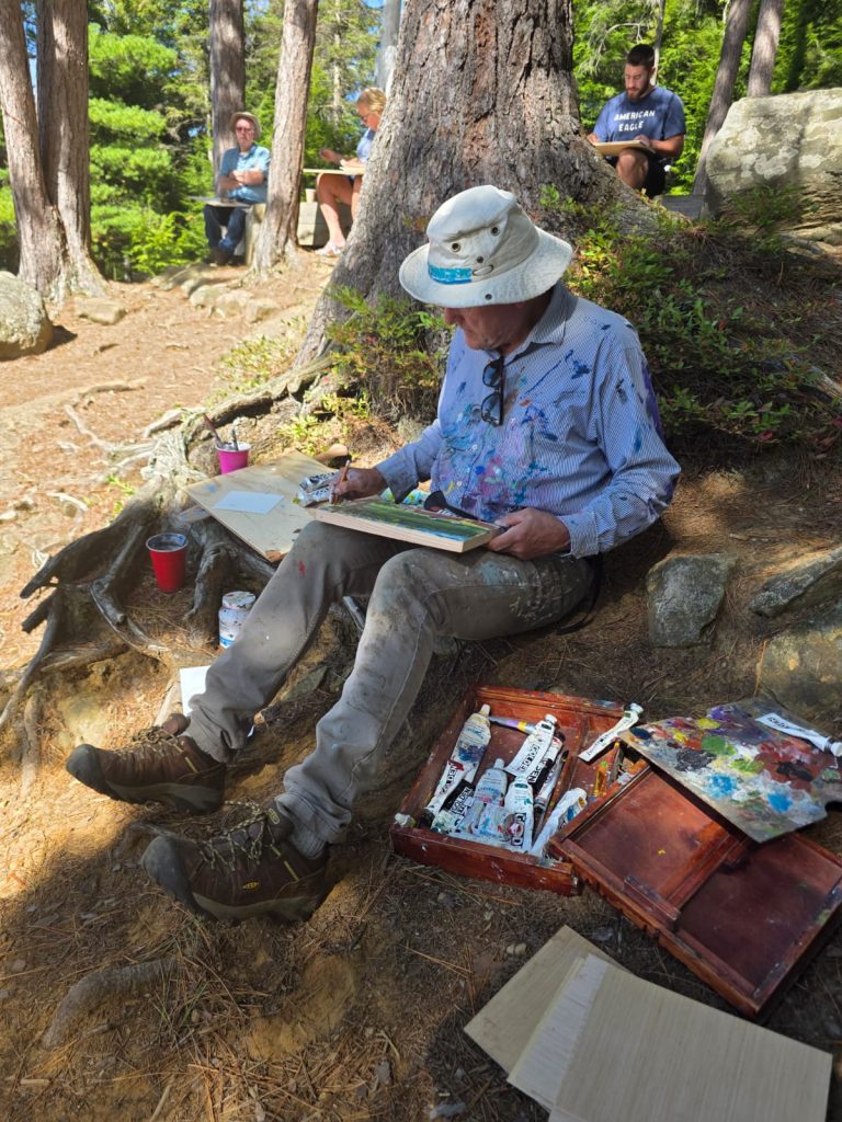A painter sits surrounded outdoors, surrounded by paint tubes and canvases