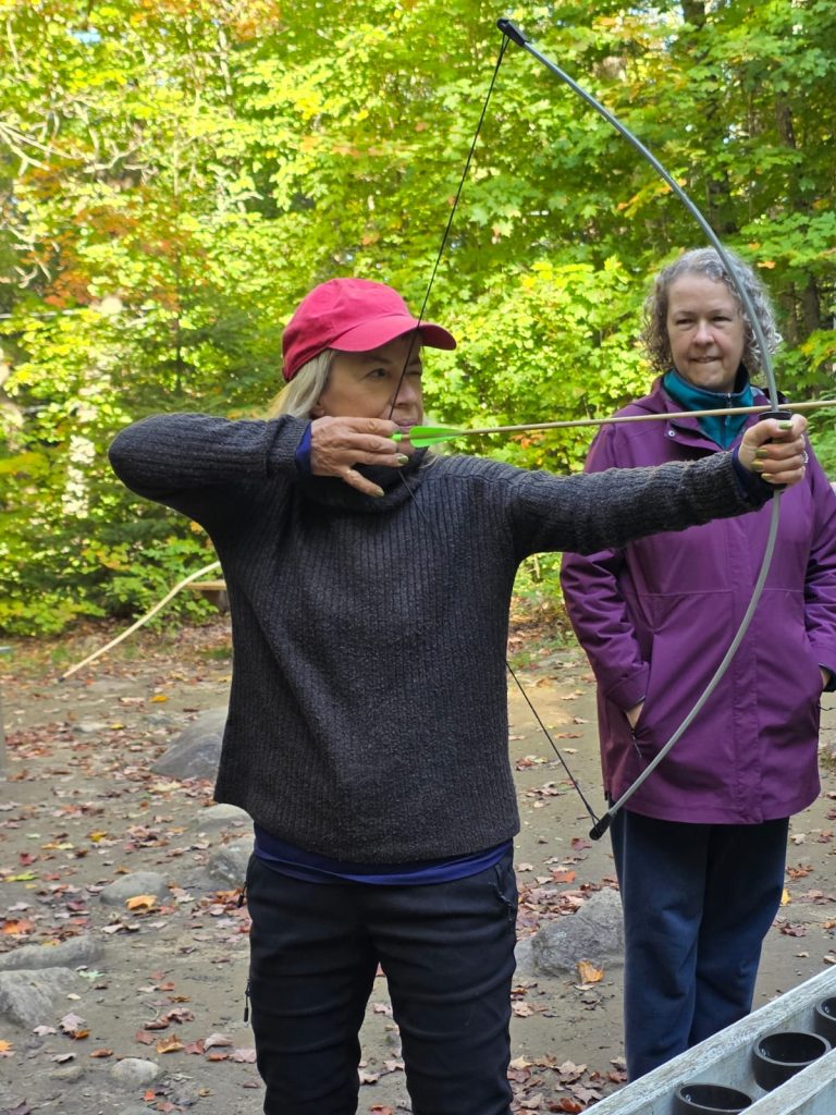 A woman draws back a bow at an archery range, while a second person looks on